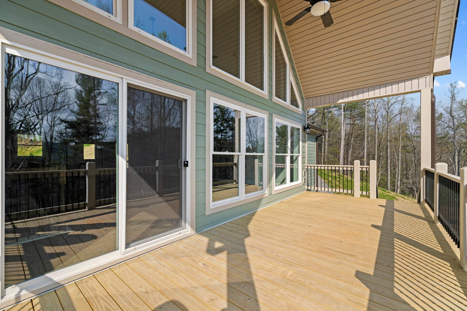 Wooden deck with screen door, ceiling fan, railing, and window; fenced yard with grassy hill and trees in background