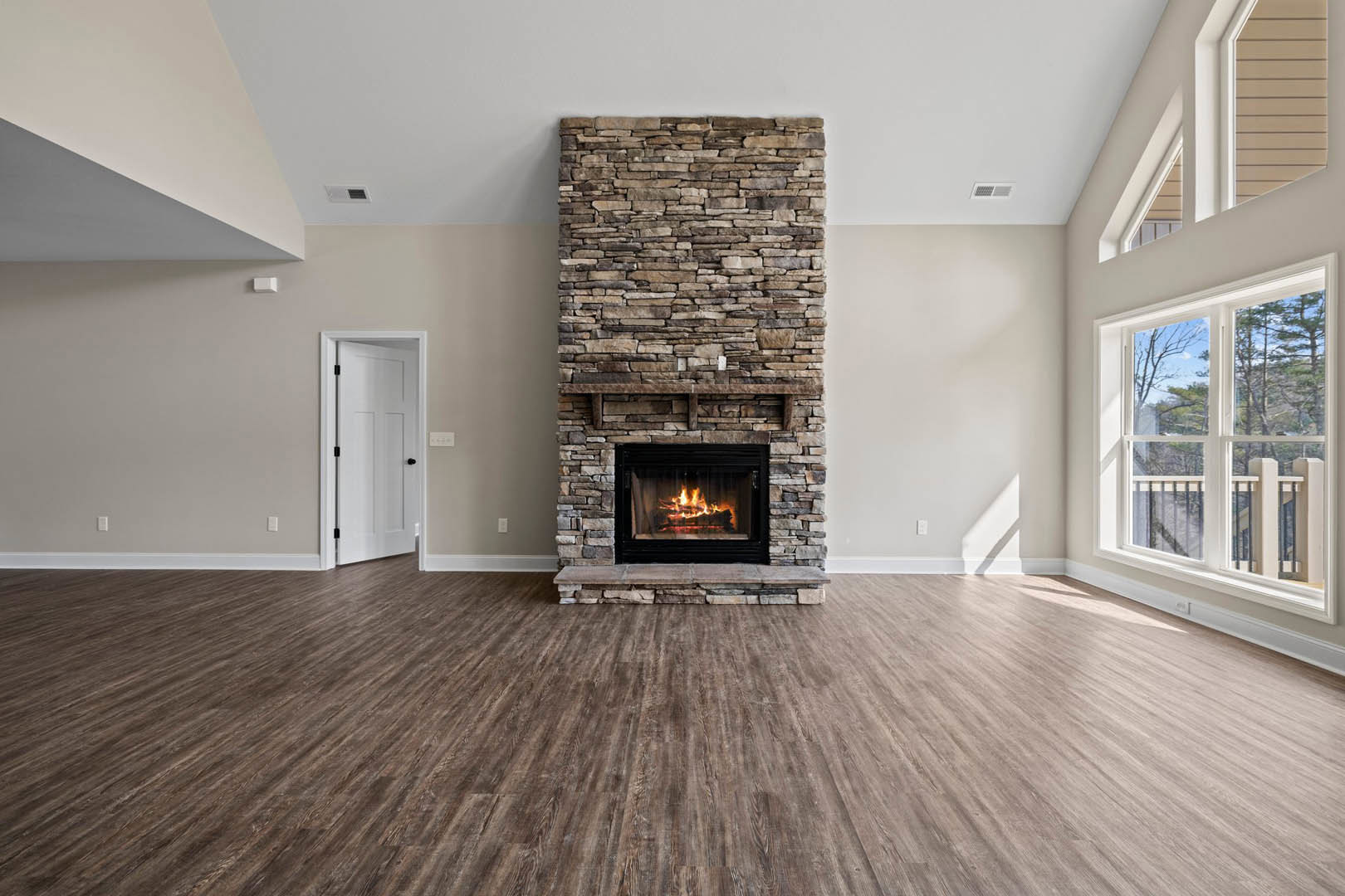 Stone fireplace with active fire, wood flooring, white door with black knobs, window featuring railing and view of trees outside