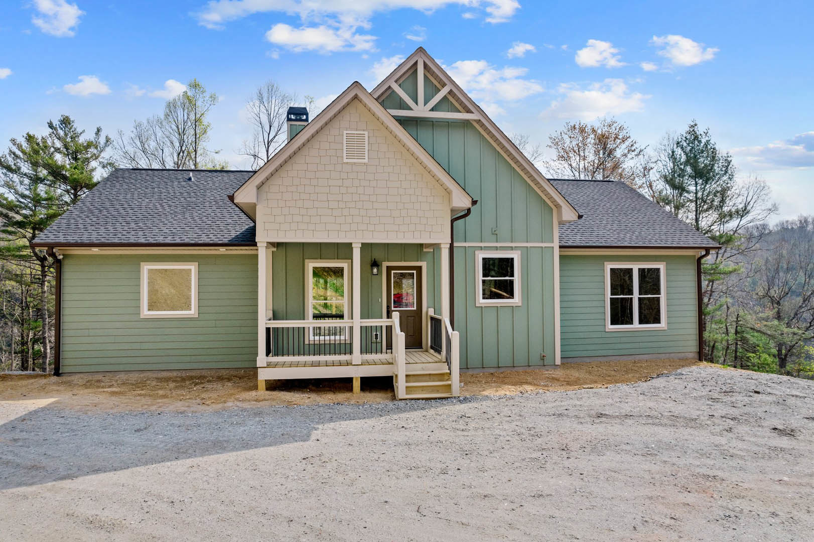 Two-story home with gray siding, white trim, and large windows, gravel driveway leading to front porch, mature trees and cloudy sky in background