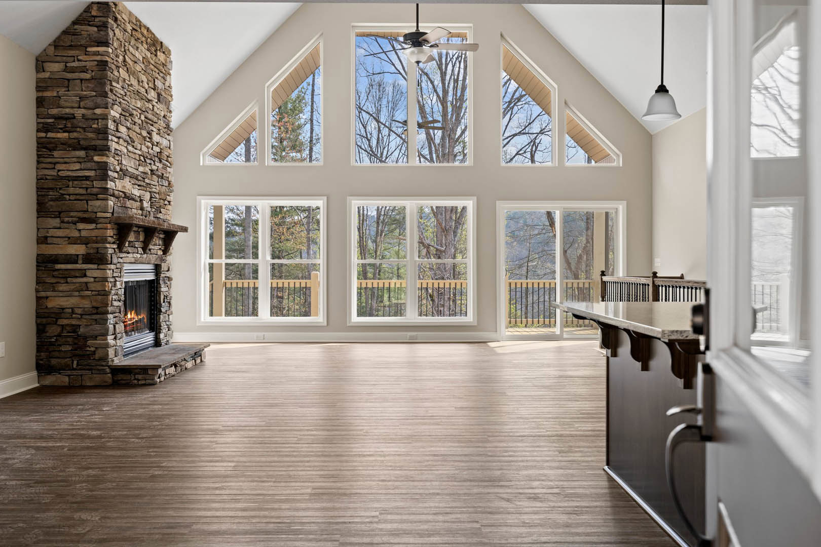 Spacious living room featuring a stone fireplace with mantel, wood flooring, ceiling fan, and large windows providing natural daylight