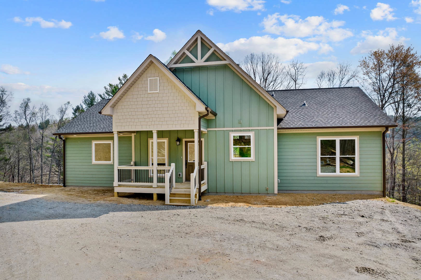 Green siding house with white porch and railing, white-trimmed windows, brick foundation, surrounded by trees and cloudy sky.
