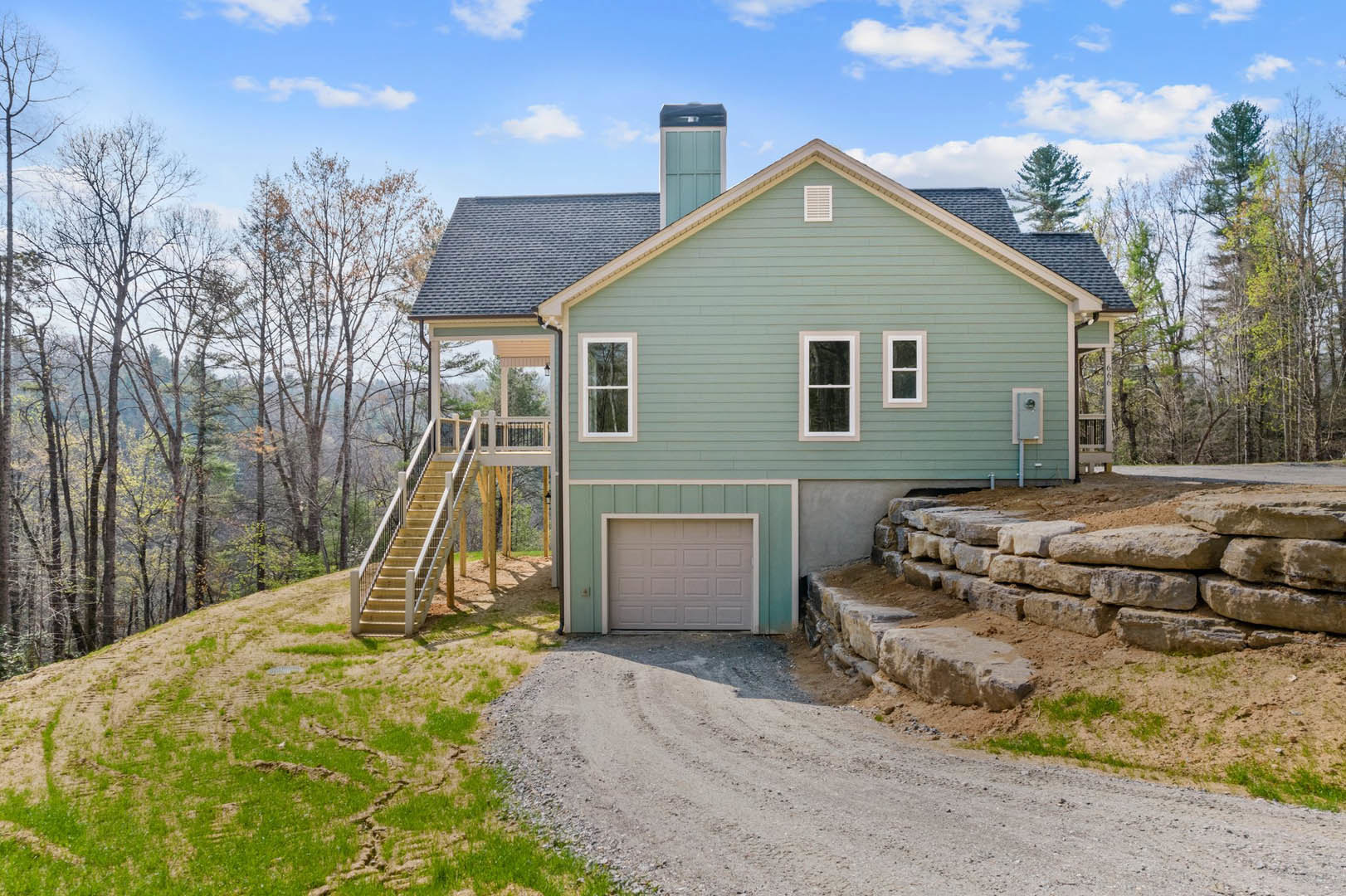 Two-story home with white garage door, green exterior wall, stone accents, white-framed windows, driveway, sloped grassy hill, and staircase leading to mature tree