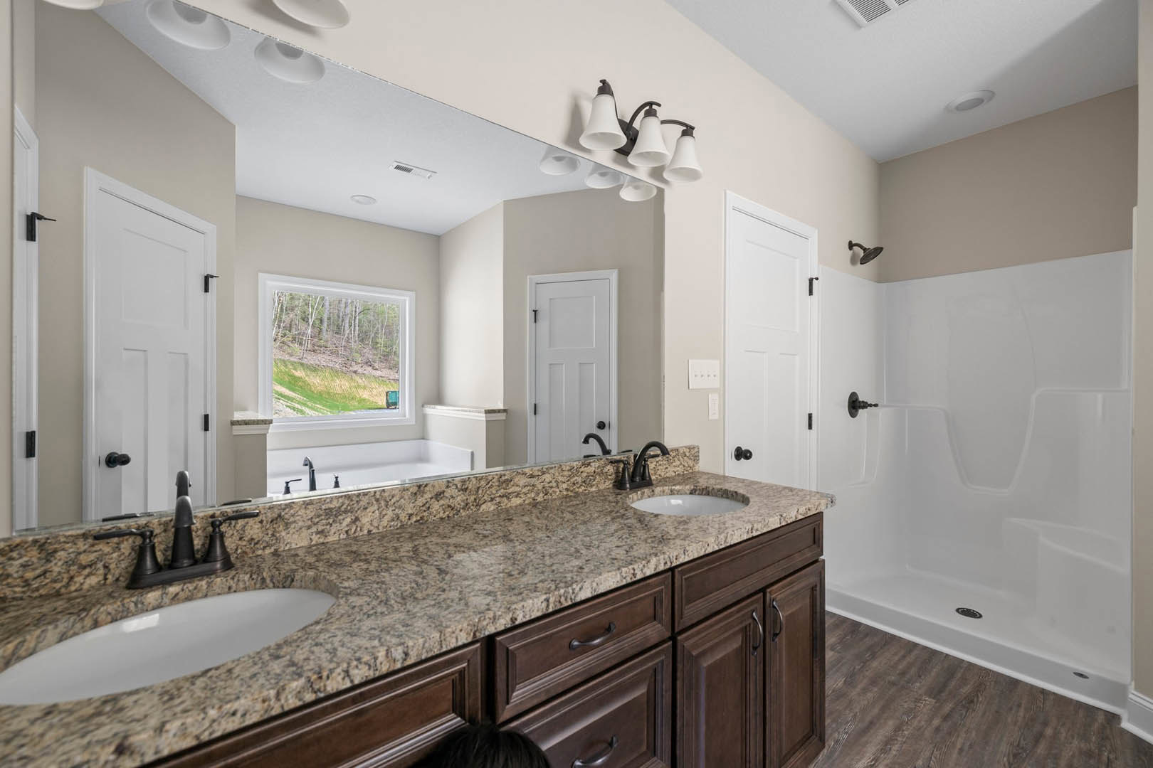 Bathroom with double sinks set in a white countertop, expansive mirror above, modern light fixture, white cabinetry with black handles, window overlooking forested landscape, light