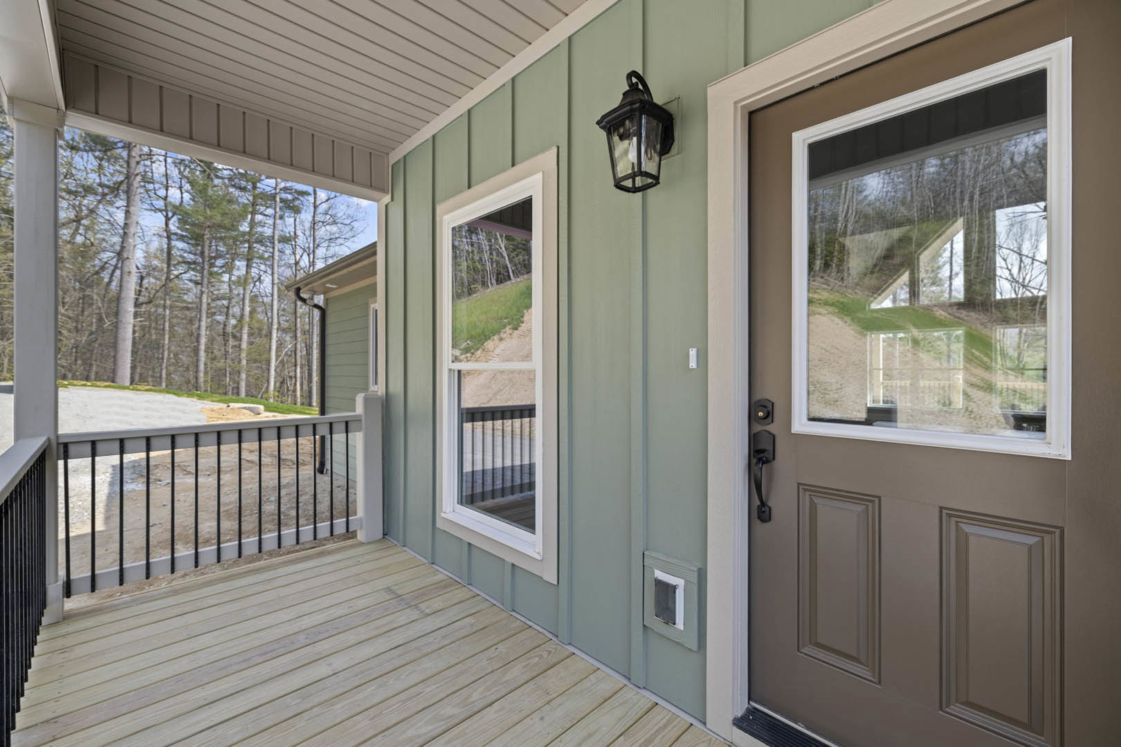 Wooden porch with black metal railing, white framed window, brown front door, and outdoor lamp fixture