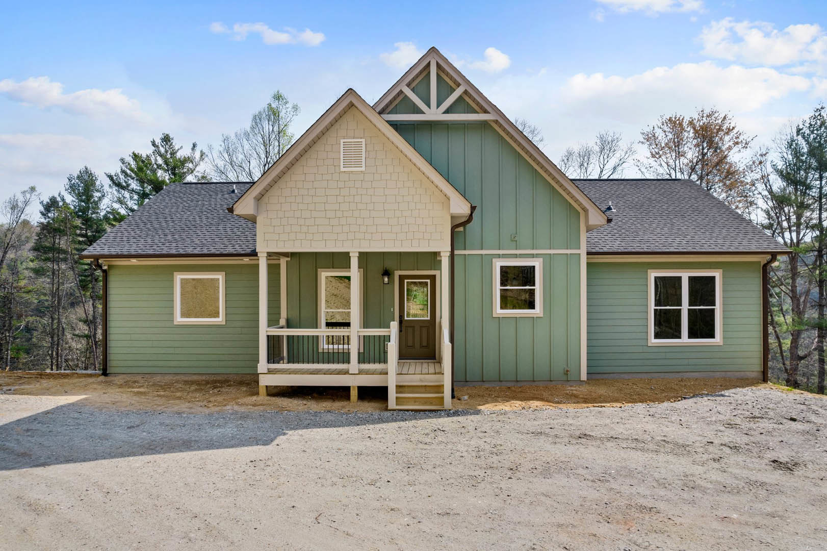 Two-story house with gravel driveway, white-framed windows, white wall vent, wooden bench on covered porch, surrounded by trees under cloudy sky
