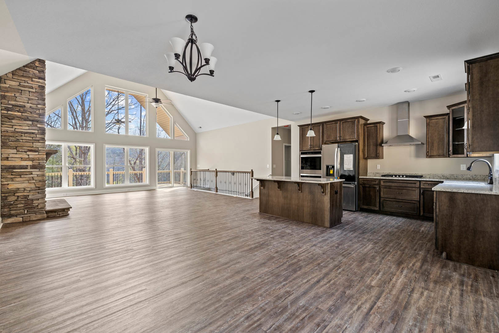Spacious open-concept kitchen with central island, wood flooring, white cabinetry, stone accent wall, modern chandelier, and stainless steel oven