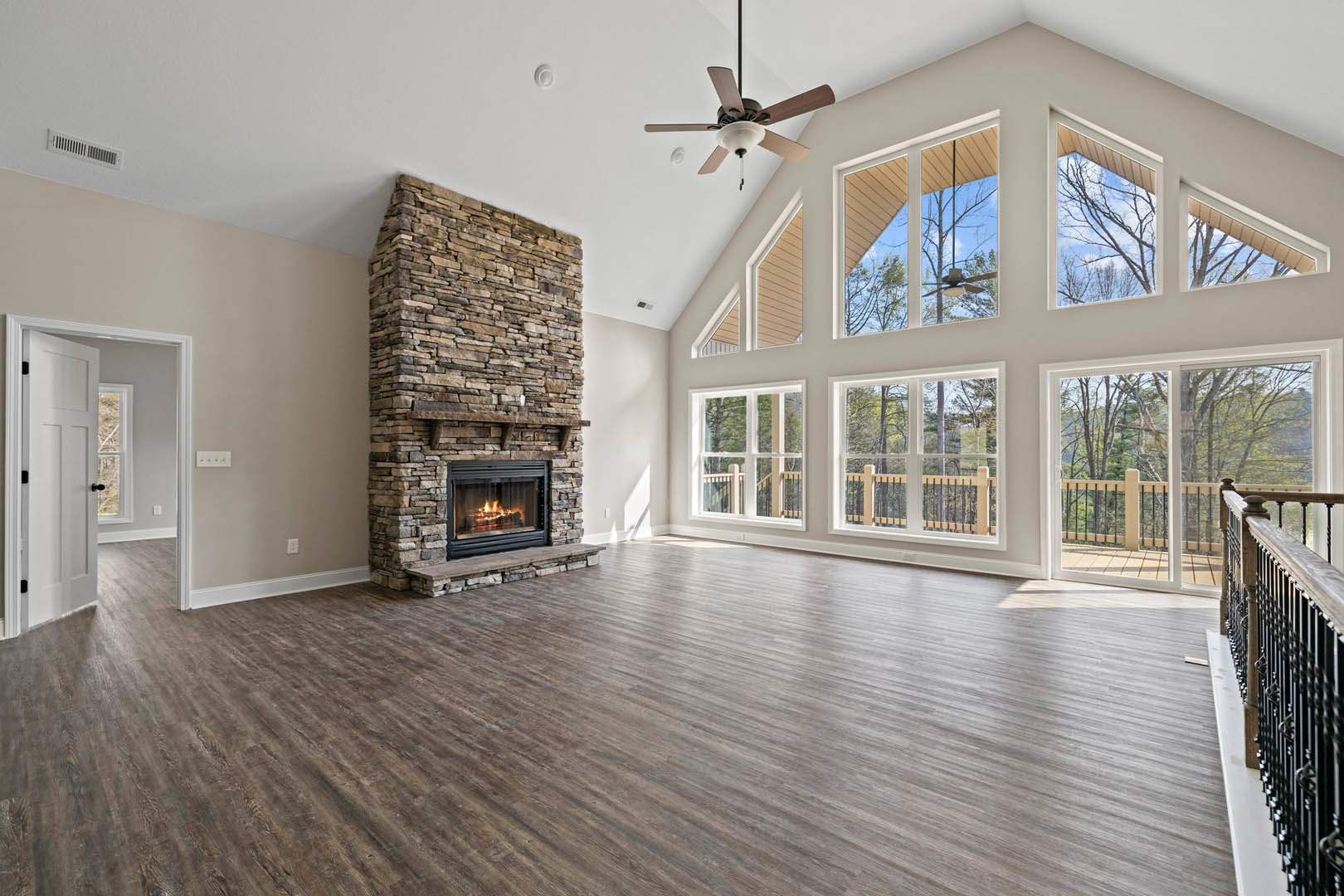 Large living room with hardwood floors, stone fireplace with fire, ceiling fan with light fixture, neutral walls