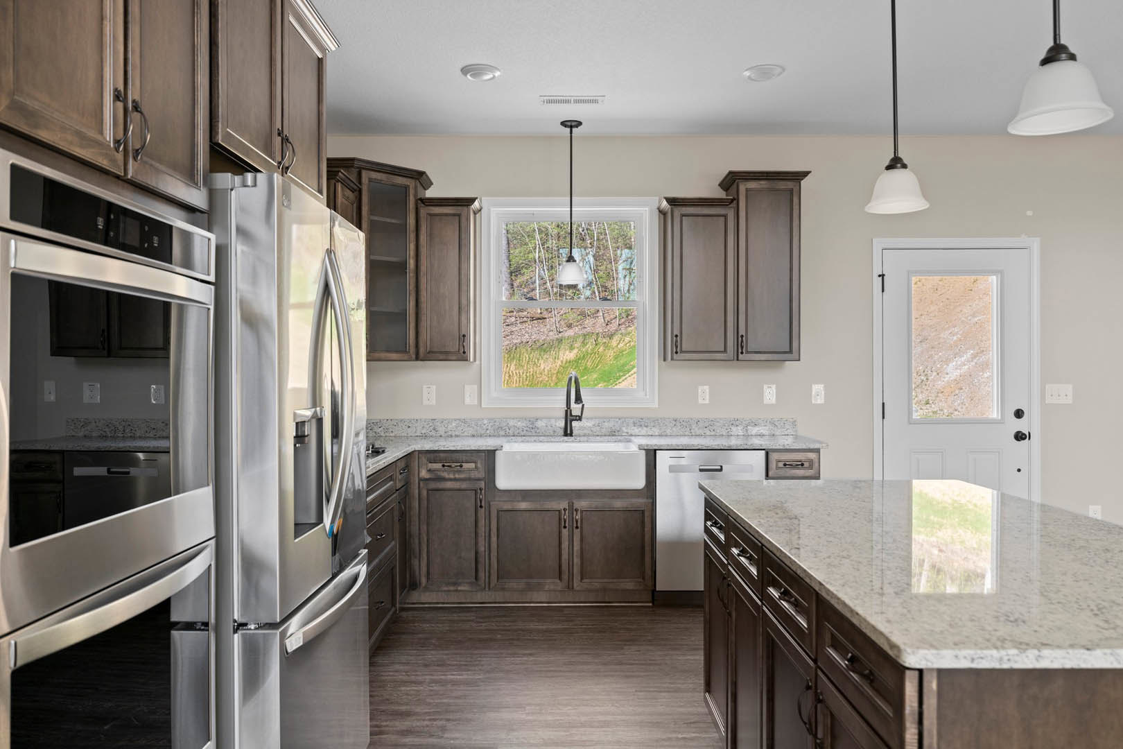 Kitchen with marble island countertop, stainless steel refrigerator with open door, wood flooring, white cabinetry, sink beneath window overlooking forest, white door with glass