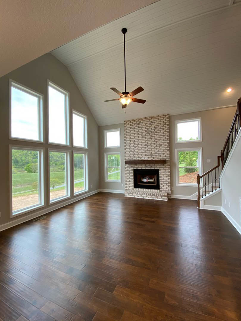 Living room with brick fireplace, ceiling fan with wooden blades and light, hardwood flooring, large windows, and decorative bottle inside fireplace