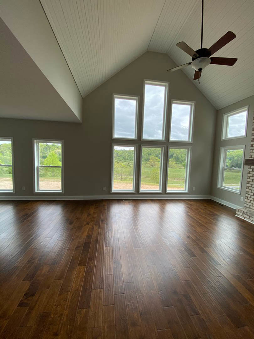 Hardwood flooring, ceiling fan with light fixture, multiple windows showing trees and cloudy sky, white walls, natural light filling the room
