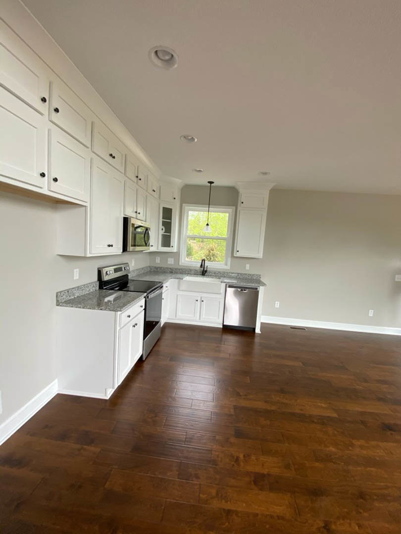 White kitchen cabinets, stainless steel refrigerator, hardwood flooring, stone countertop with built-in stove and dishwasher, window letting in natural light