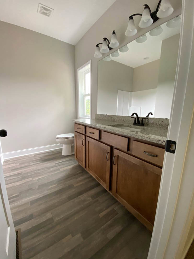 Bathroom with wood flooring, white toilet, rectangular sink on a stone countertop, large mirror with row of lights above, white ceiling and wall vents.
