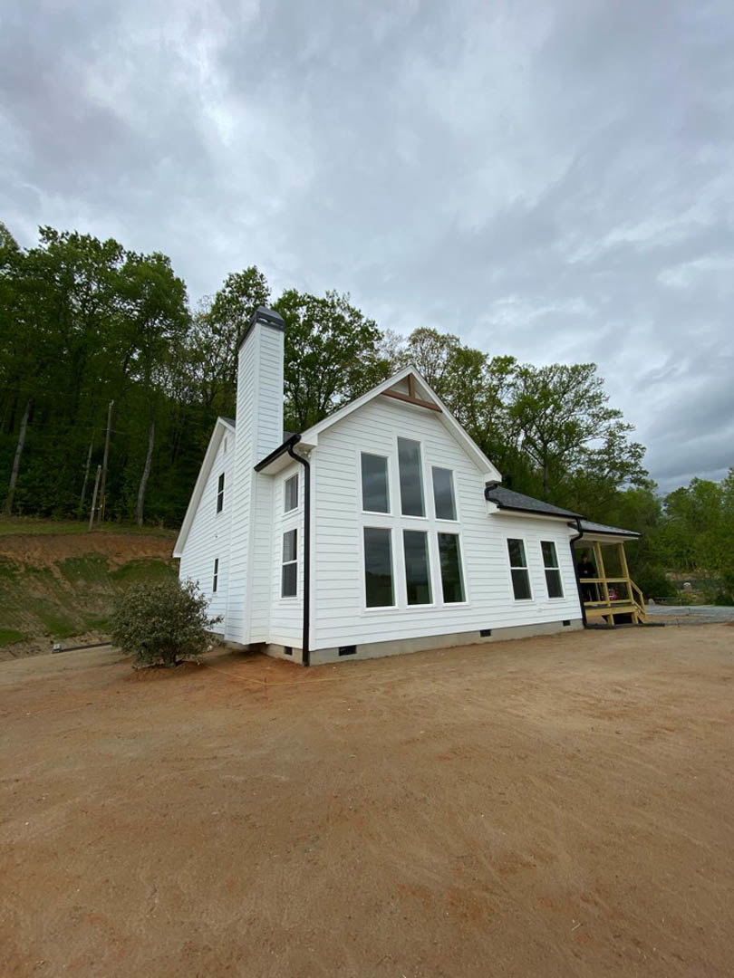 White cottage-style home with brick chimney, large windows, covered porch, mature trees in background, dirt driveway in foreground, person standing on porch