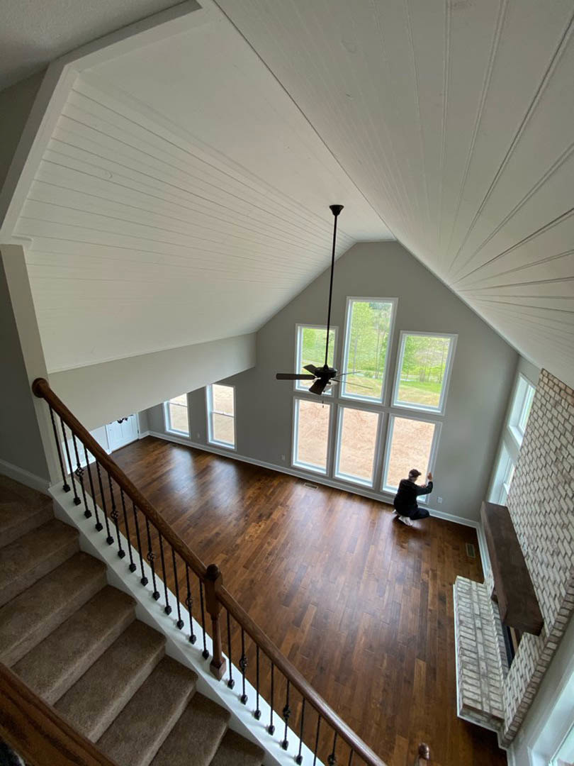 Open living area with carpeted staircase, hardwood floors, large windows, ceiling fan, and a person seated on an upholstered chair