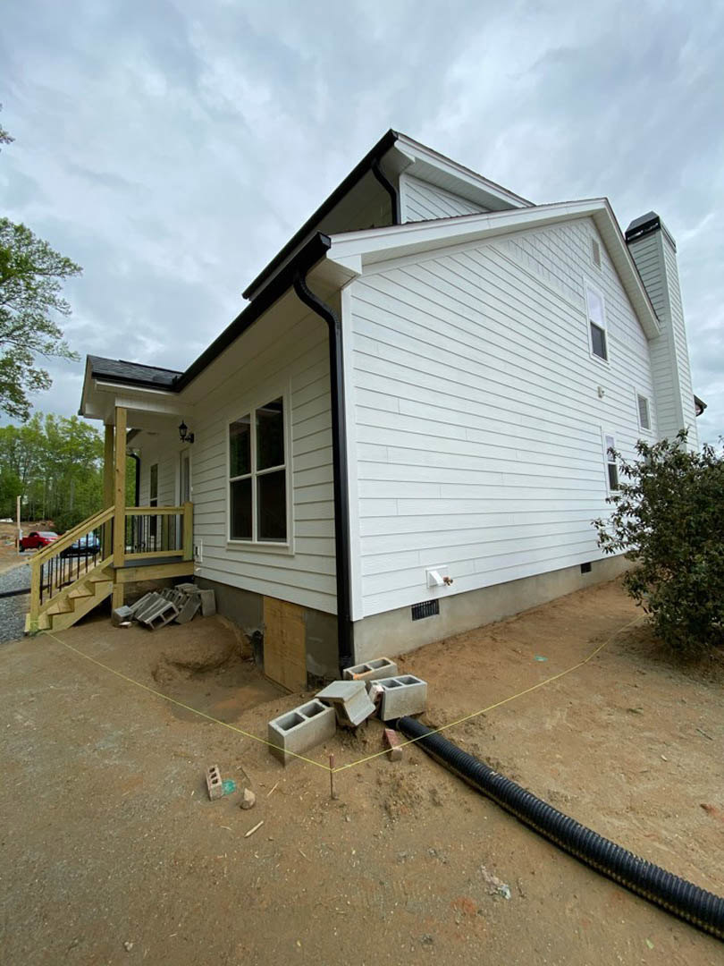 Partially built home with white framed windows, unfinished siding, covered porch, black pipe lying on bare ground, leafy tree and bush in front yard under cloudy sky