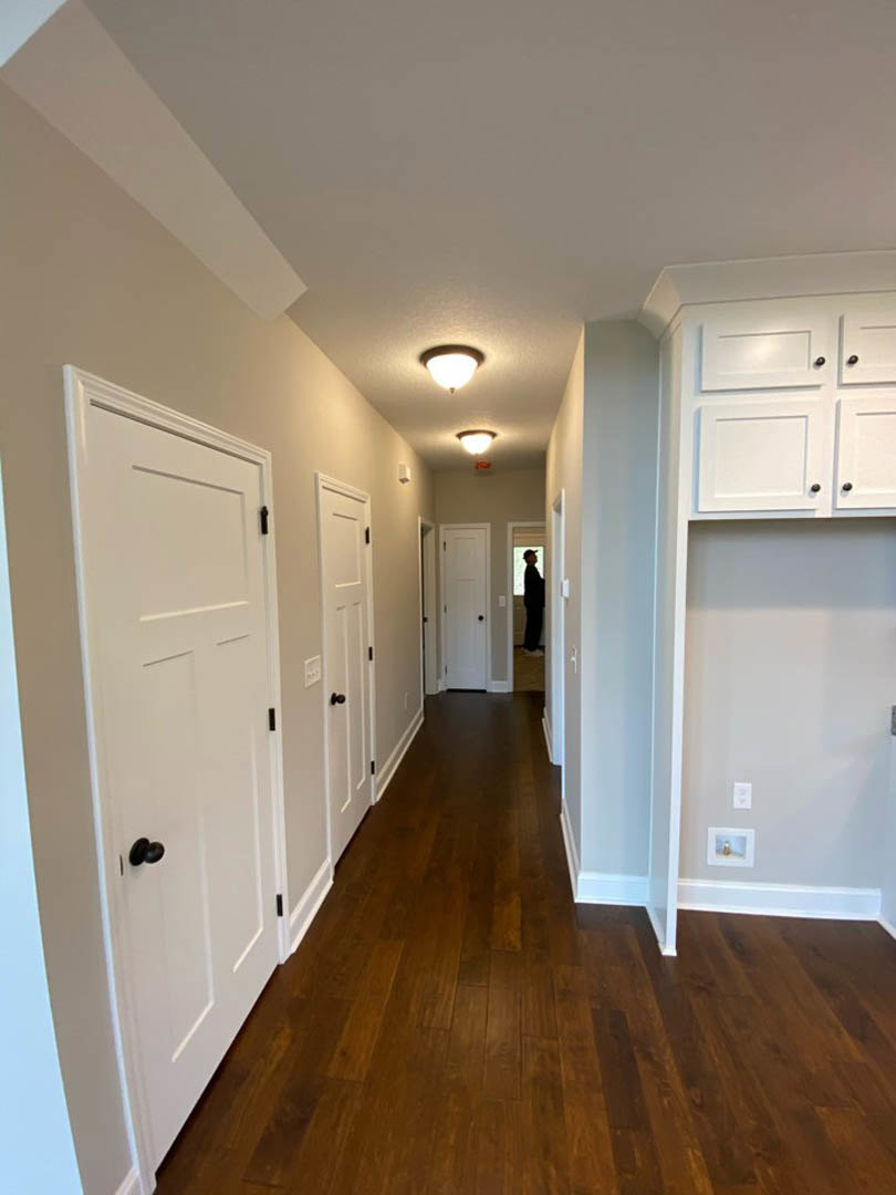 Hallway with white paneled doors, dark wood laminate flooring, white cabinetry with black knobs, ceiling light fixture, and white walls