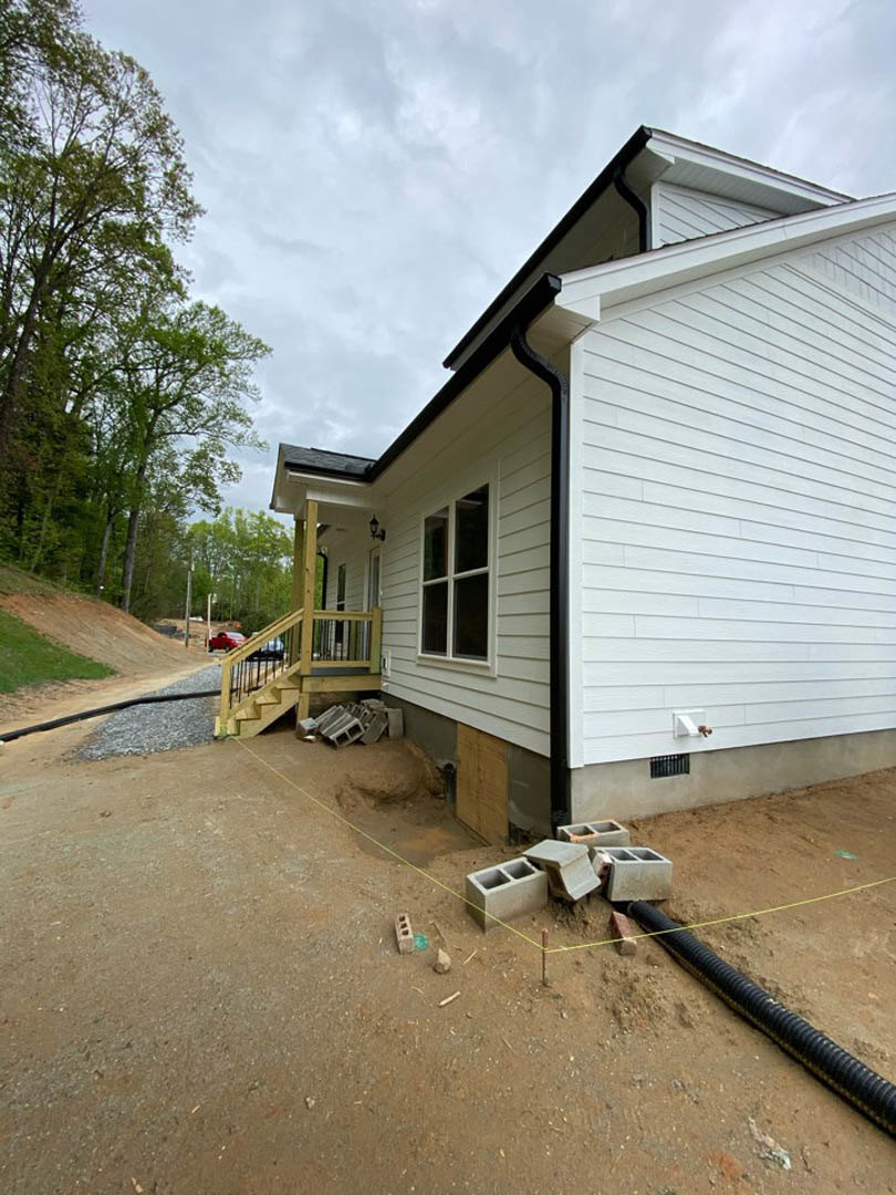 White house under construction with exposed staircase, white-framed window, black pipe and construction materials scattered on bare ground, bordered by a path and road, surrounded