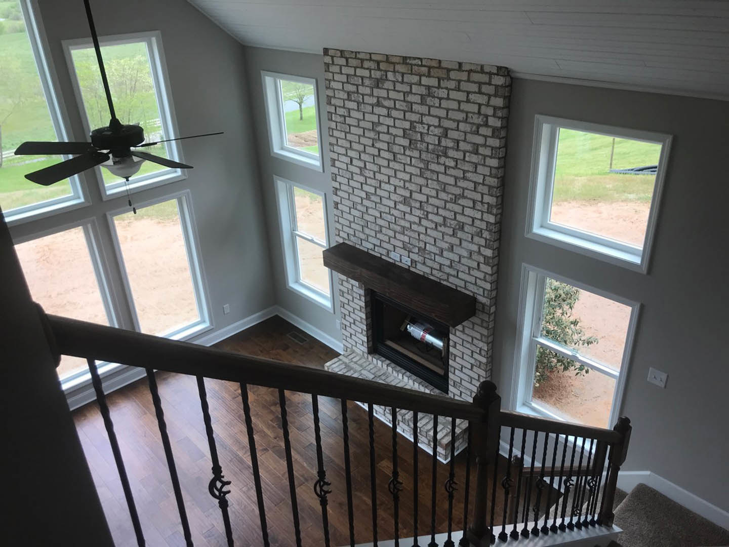 Wood staircase with iron balusters overlooking living room fireplace, white walls, ceiling fan, and window blinds.