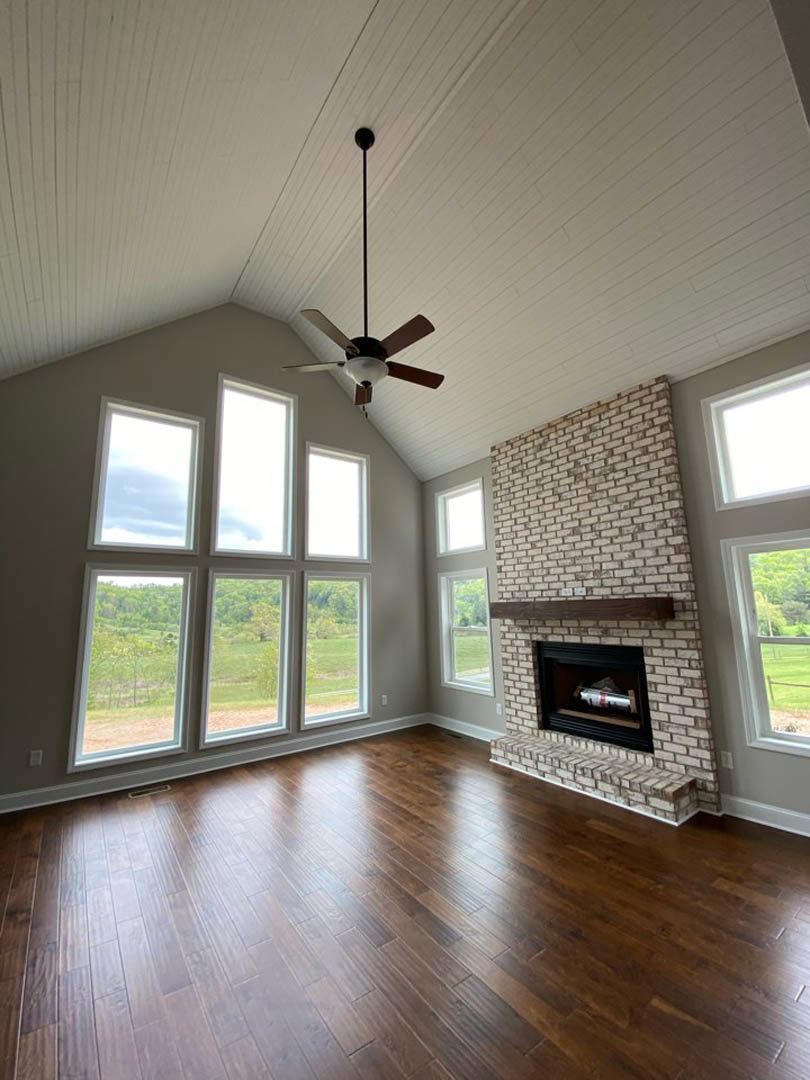 Hardwood floored living room with brick fireplace, wood mantel, silver bottle decor, ceiling fan with light, and wall of multiple windows