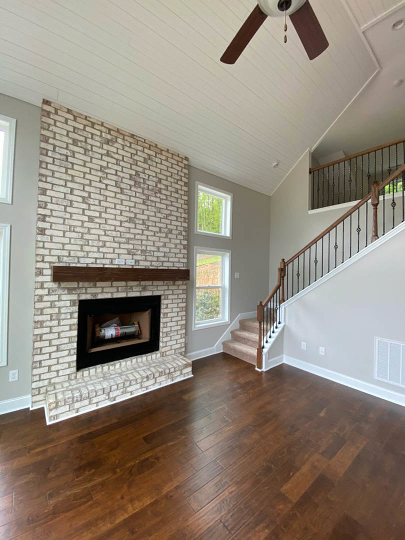 White brick fireplace with black rectangular hearth, hardwood floors, window overlooking grassy field, and staircase with wood treads.