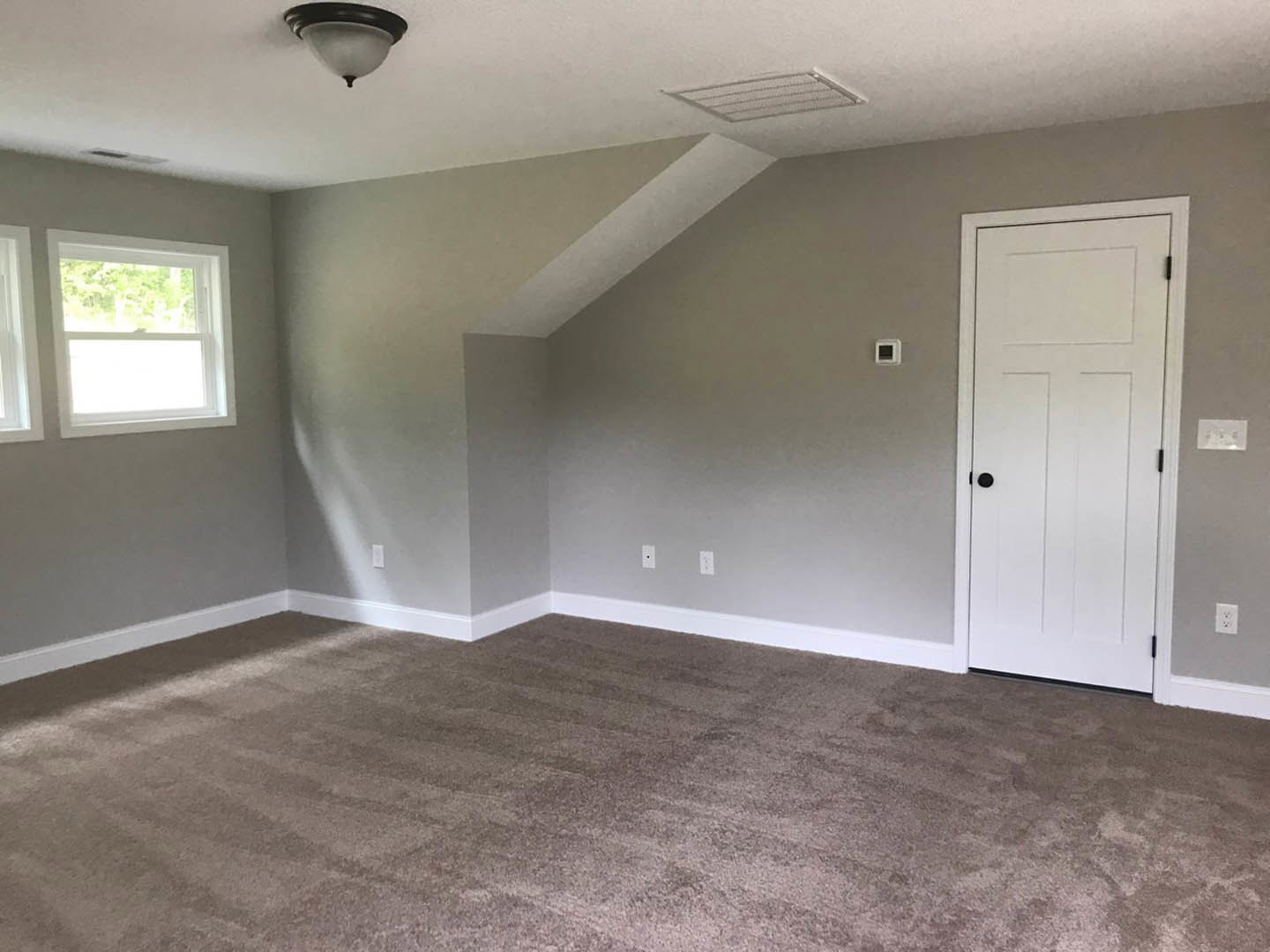 White paneled door with black knob, window with white frame, beige carpeted floor, white walls, ceiling light fixture, white electrical outlet with black text