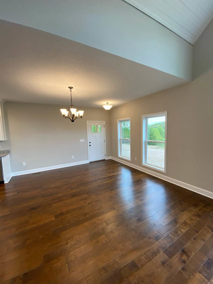 Room with hardwood floor, two large windows overlooking trees, white door with glass panel, and modern chandelier hanging from ceiling