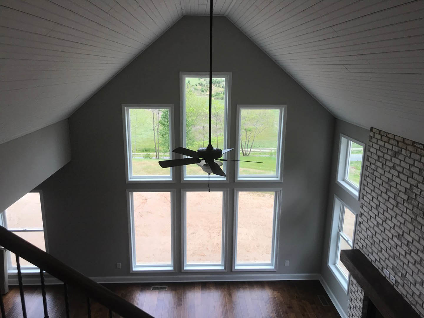 Ceiling fan mounted in a bright room with large windows, brick fireplace, hardwood floors, and staircase with wooden railing