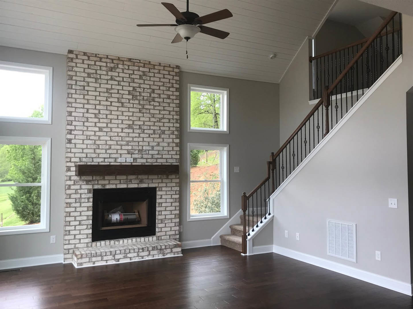 Living room featuring a stone fireplace, wooden staircase with white risers, ceiling fan with light, large windows showing outdoor landscape, hardwood floors, and neutral wall