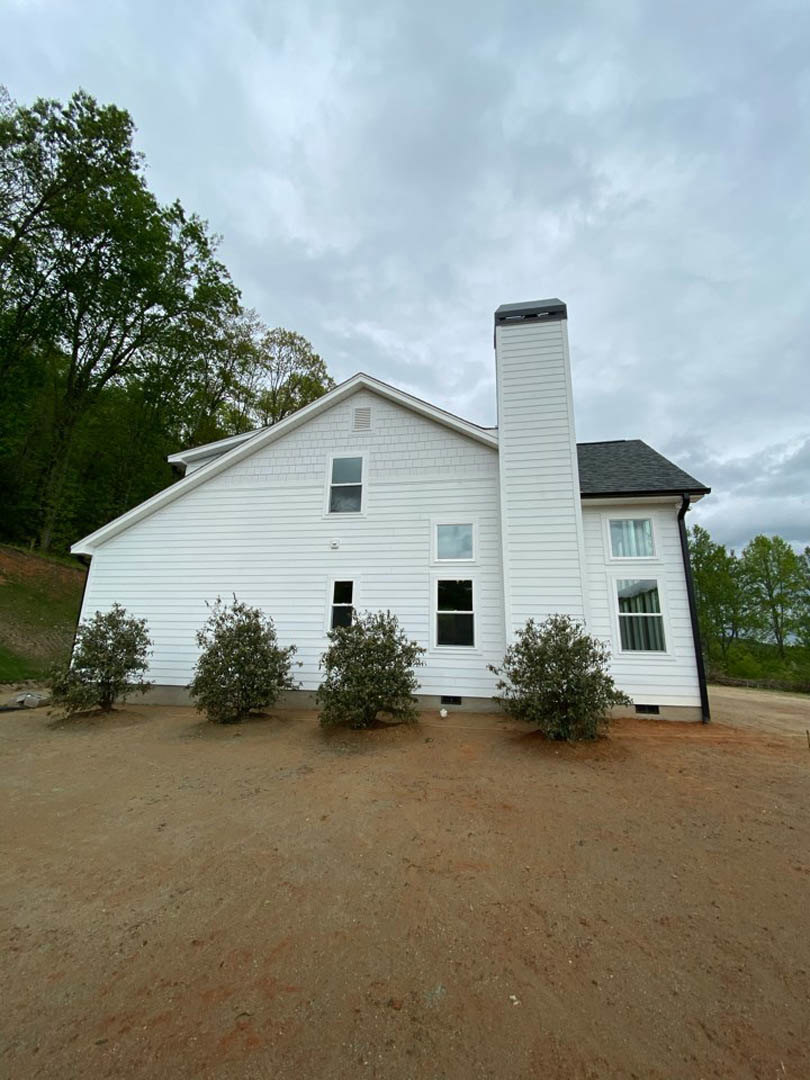 White cottage-style home with brick chimney, gabled roof, and large leafy trees surrounding the property; windows visible on the front facade, dirt field in foreground.