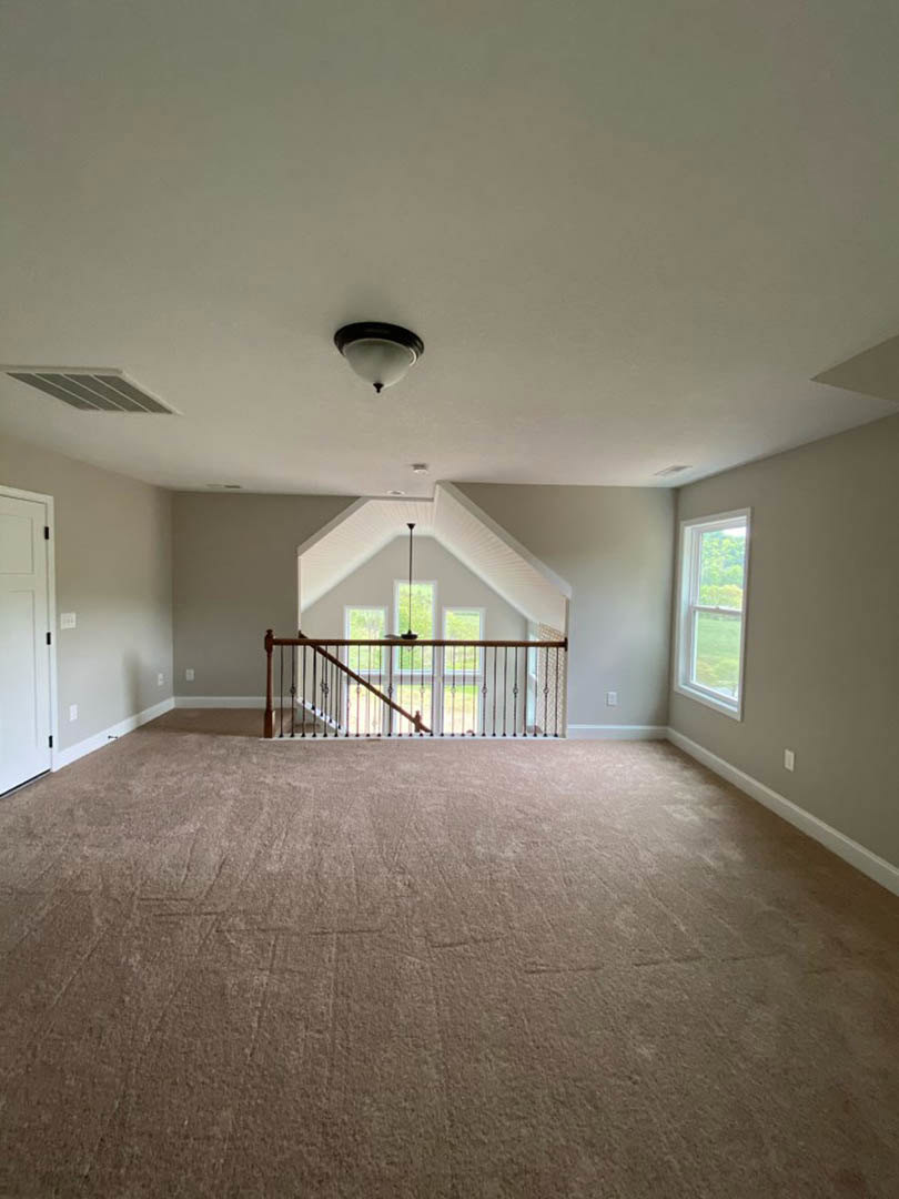 Open living area featuring a carpeted floor, wooden staircase with metal railing, large window overlooking a green field, and ceiling light fixture.