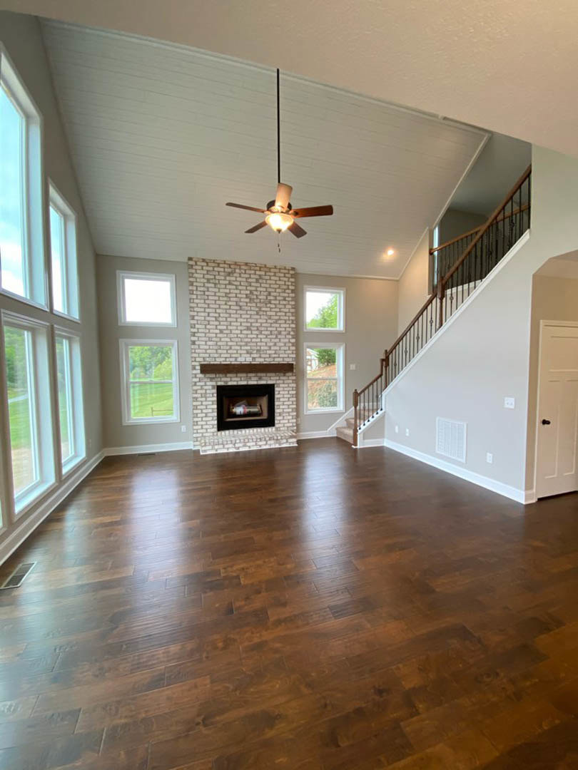Living room featuring brick fireplace with a bottle inside, hardwood floors, wood staircase railing, and ceiling fan with light fixture