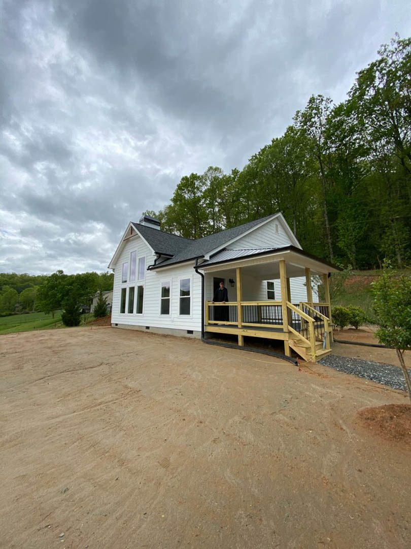 Wooden porch with black railing, stairs leading to dirt path, man standing on porch, house surrounded by trees under cloudy sky