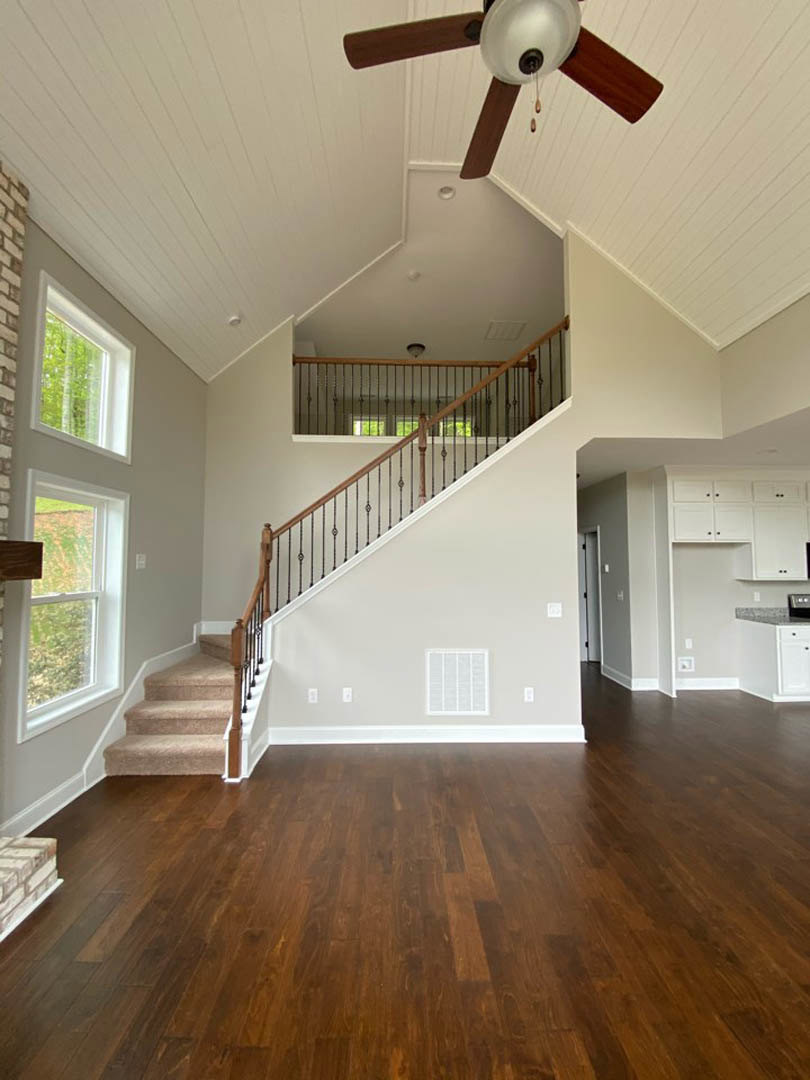 Hardwood floor living space featuring a carpeted staircase with metal railing, ceiling fan, and large window overlooking grassy field