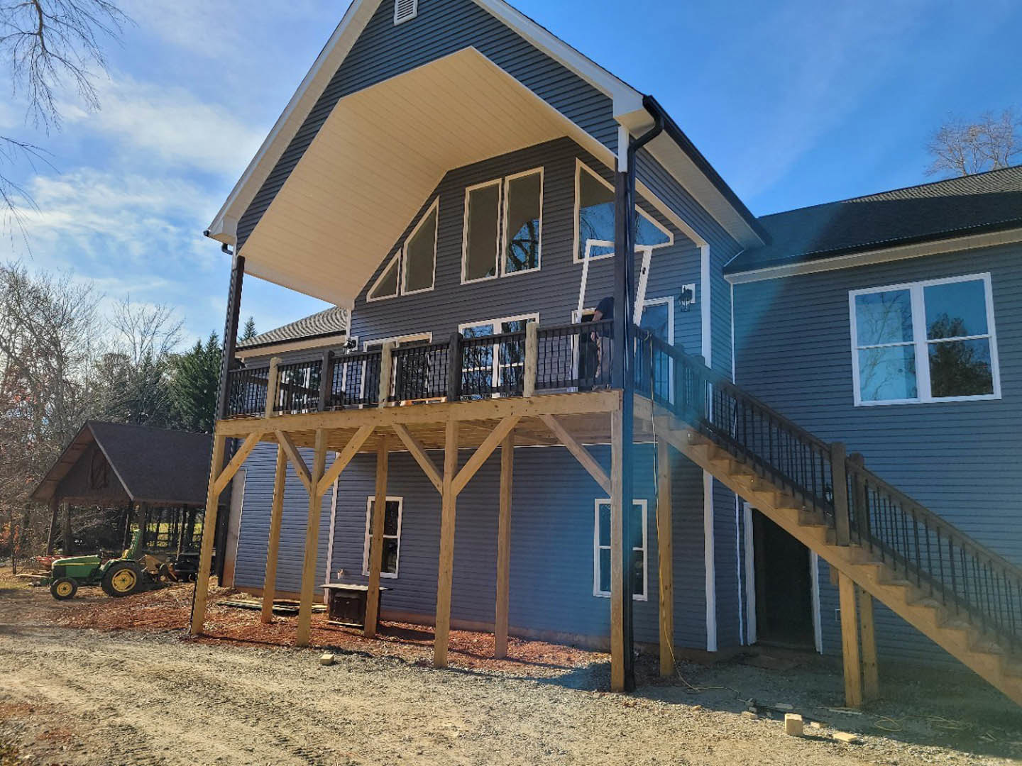 Wood deck with white stairs leading to a two-story house, white-framed windows, blue glass accents, green tractor with yellow wheels parked nearby, trees and clouds in the