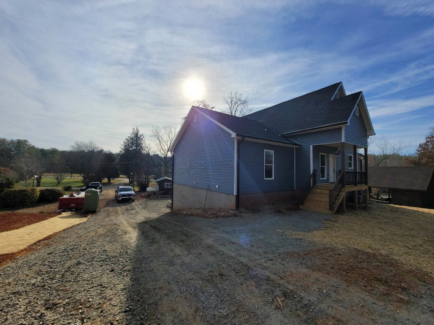 Two-story house with covered porch and stairs, concrete driveway, parked cars, green container, leafy trees, bright sun shining through scattered clouds