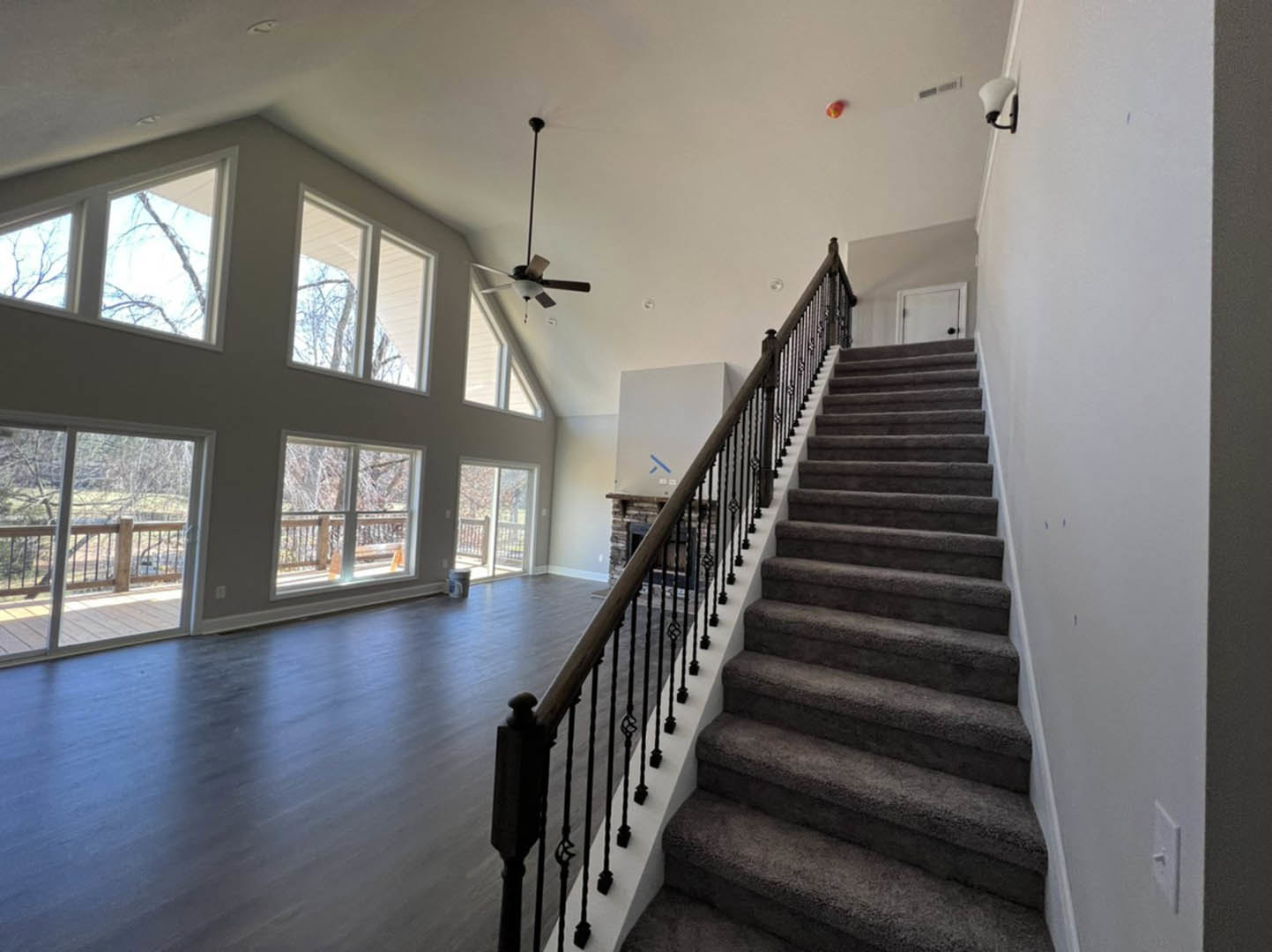 Carpeted staircase with wooden handrail in spacious room featuring large windows, white door with black knobs, and deck access; tree branches visible through window.