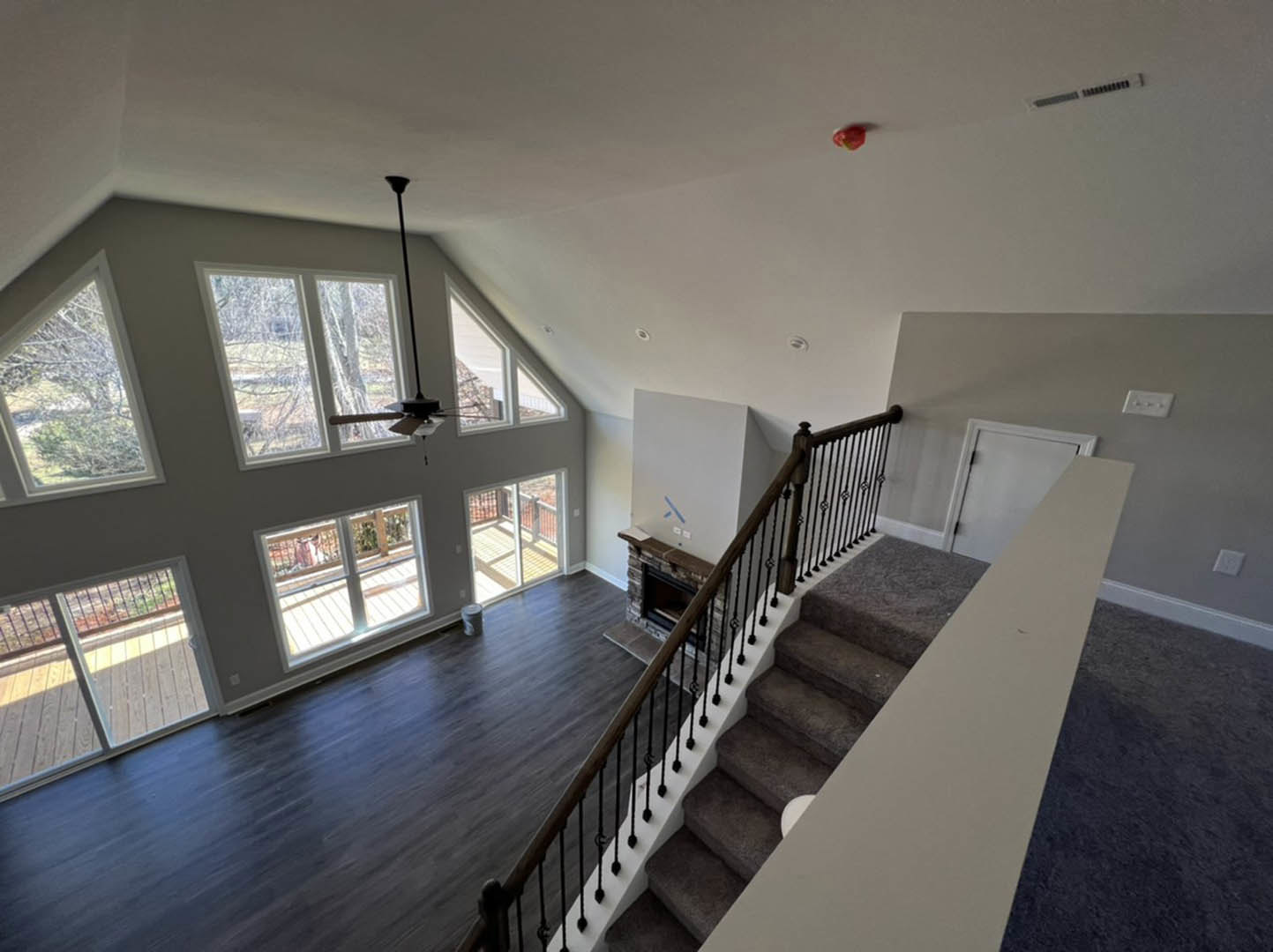 Open living room with hardwood floors, modern fireplace, ceiling fan, and staircase; sliding glass door leads to wooden deck; large windows provide natural daylight.