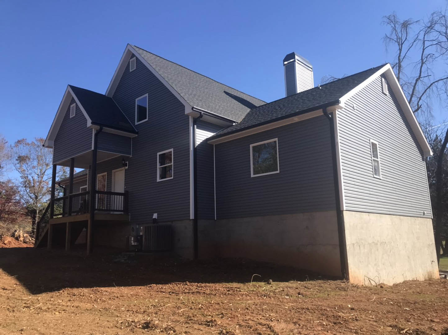 Two-story house with white siding, front porch, elevated wooden deck, white-framed windows, brick chimney, surrounded by trees and dirt yard.
