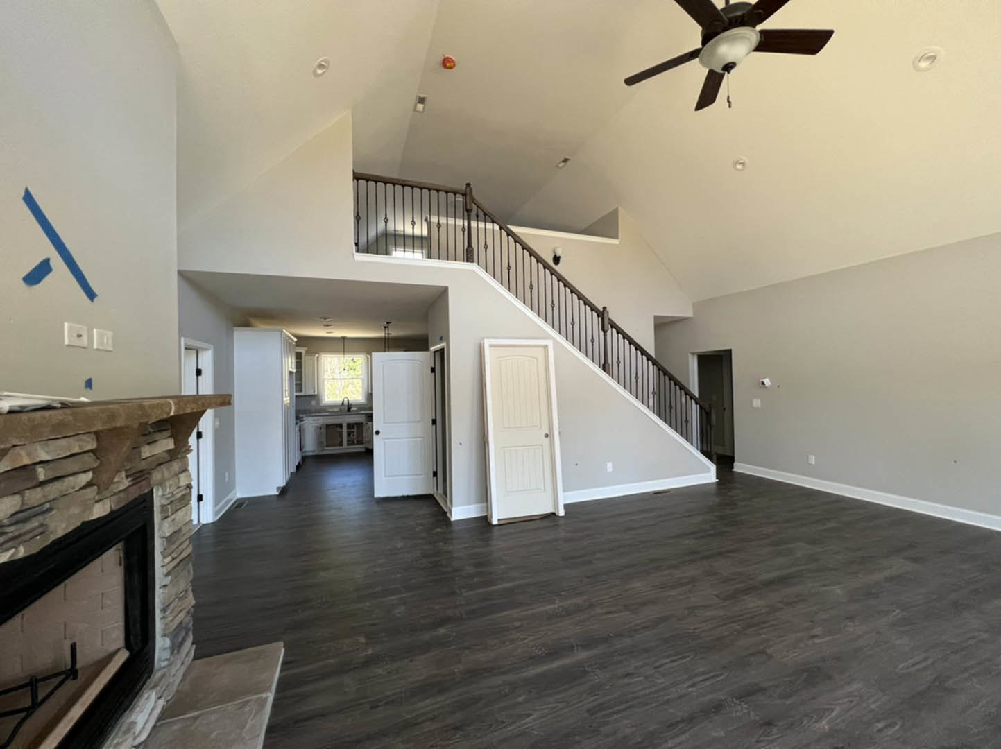 Living room with dark wood flooring, white door with black knobs, ceiling fan with light fixture, fireplace, and blue painter’s tape on white wall.