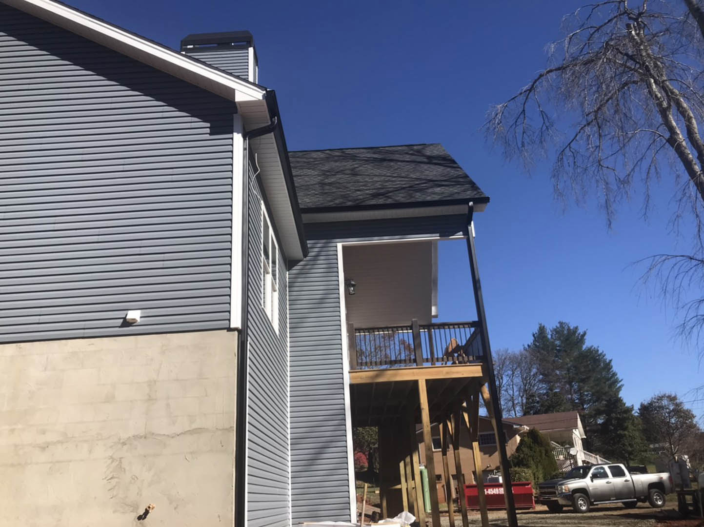 Two-story home with light siding, elevated wooden deck, leafless tree in front yard, silver pickup truck parked on dirt driveway, red and white sign near entrance, windows along