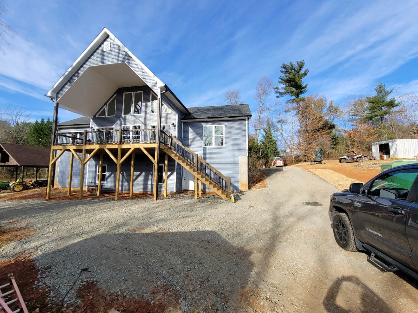 Two-story house with white siding, wooden porch and deck, black truck parked on gravel driveway, large windows with white frames, trees and cloudy sky in background