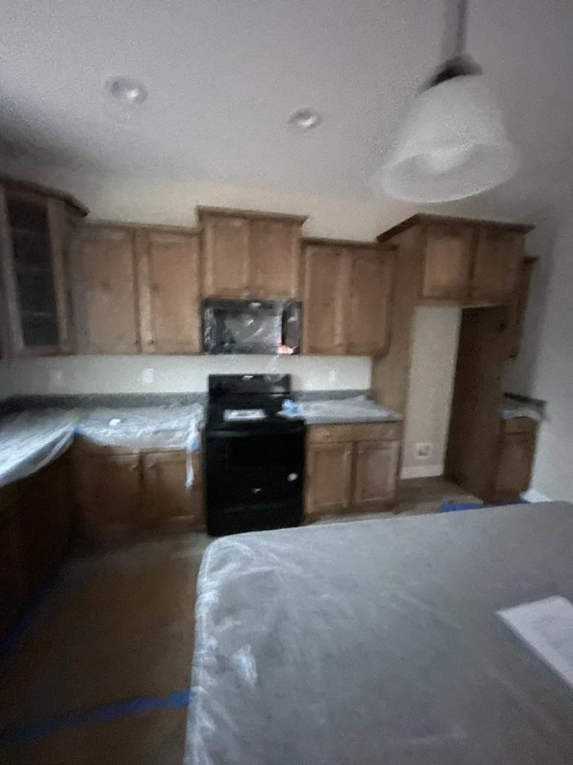 Open kitchen with light wooden cabinets, white countertop, stainless steel sink, and adjacent bedroom featuring a bed with white sheets.
