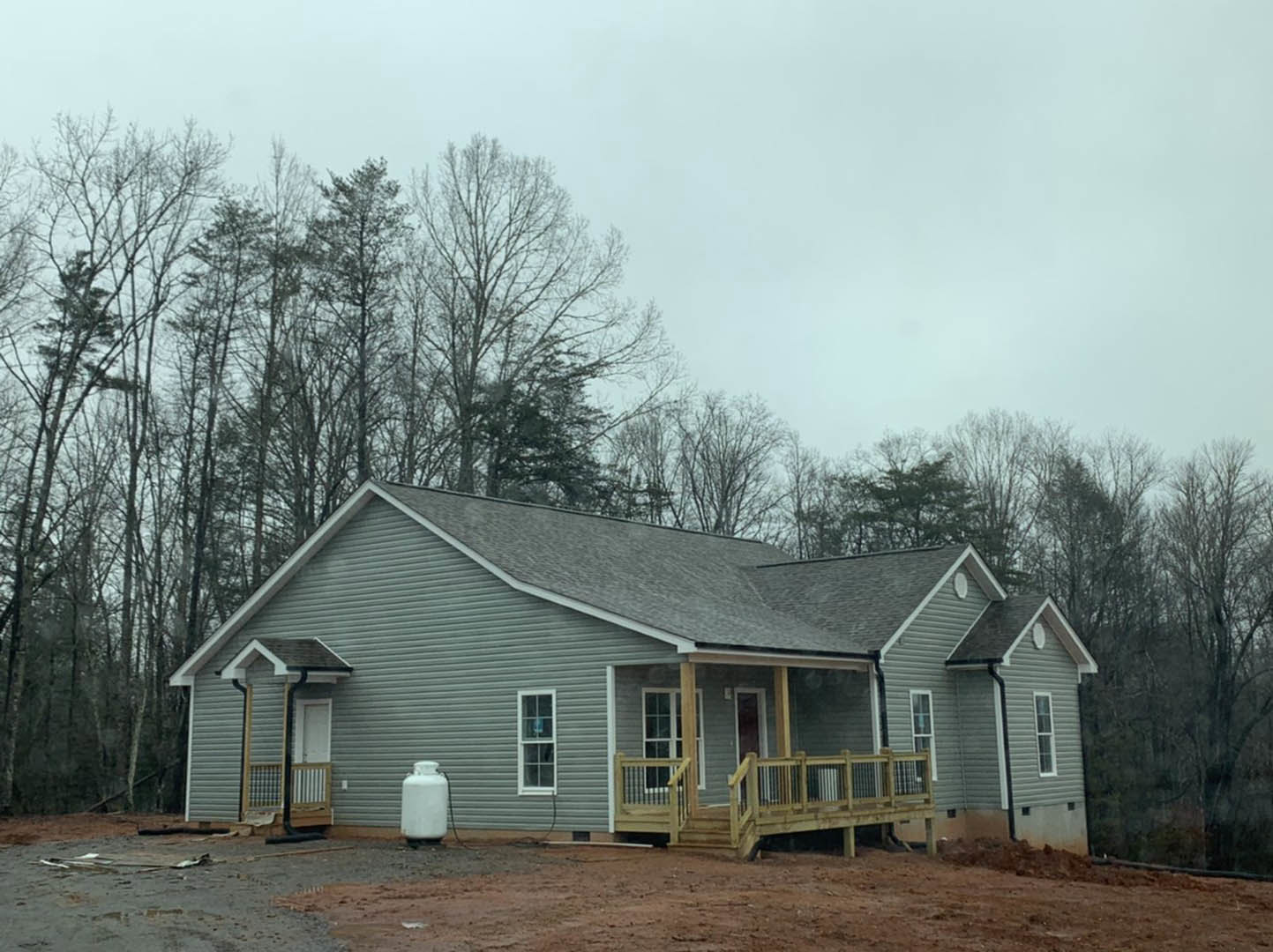Partially built house with wood deck and railing, white-framed window, exposed siding, surrounded by trees under cloudy sky