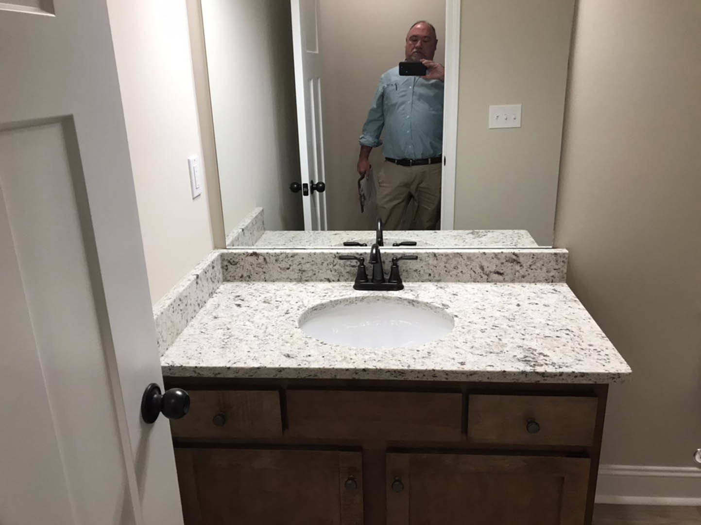 Man taking a selfie in bathroom with white tile walls, black faucet on white sink, three switches on wall, and close-up of silver door knob
