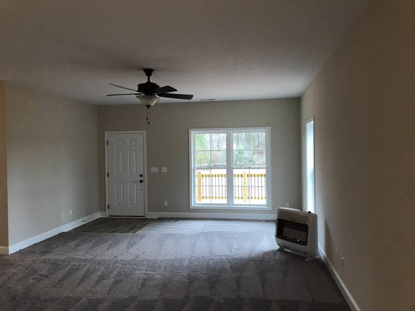 Neutral-toned carpeted room with a ceiling fan and light, large window showing outdoor view, white door with black hardware, and floor heater along the wall