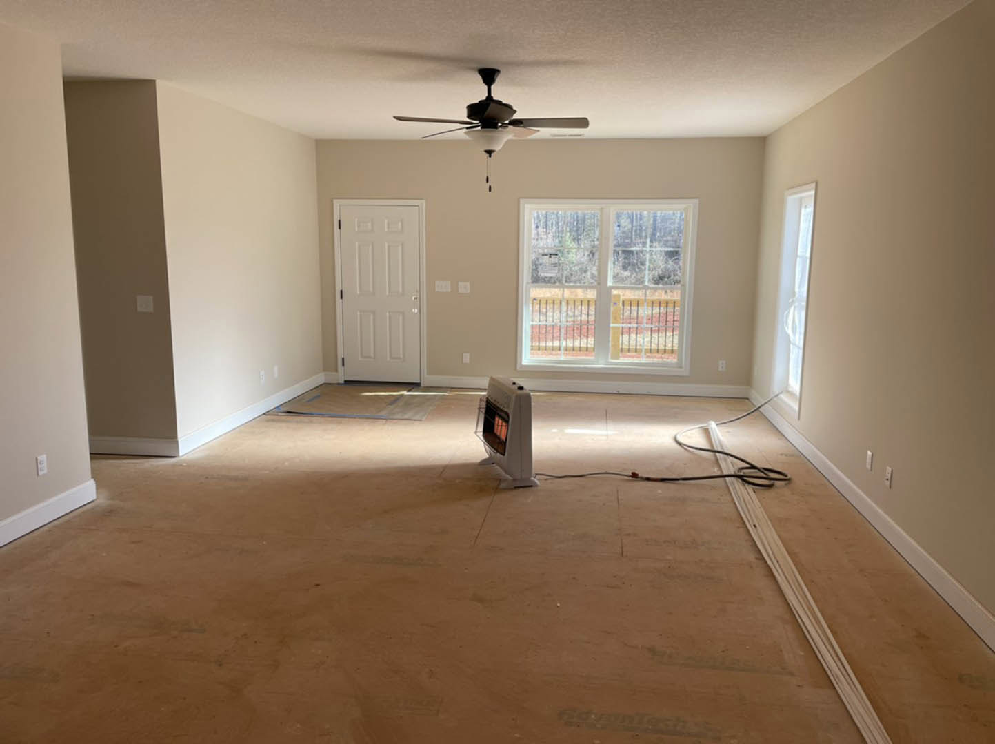 Laminate wood flooring, white plaster walls, ceiling fan with light, wall-mounted heater, window overlooking fenced yard and trees, white door with metal doorknob