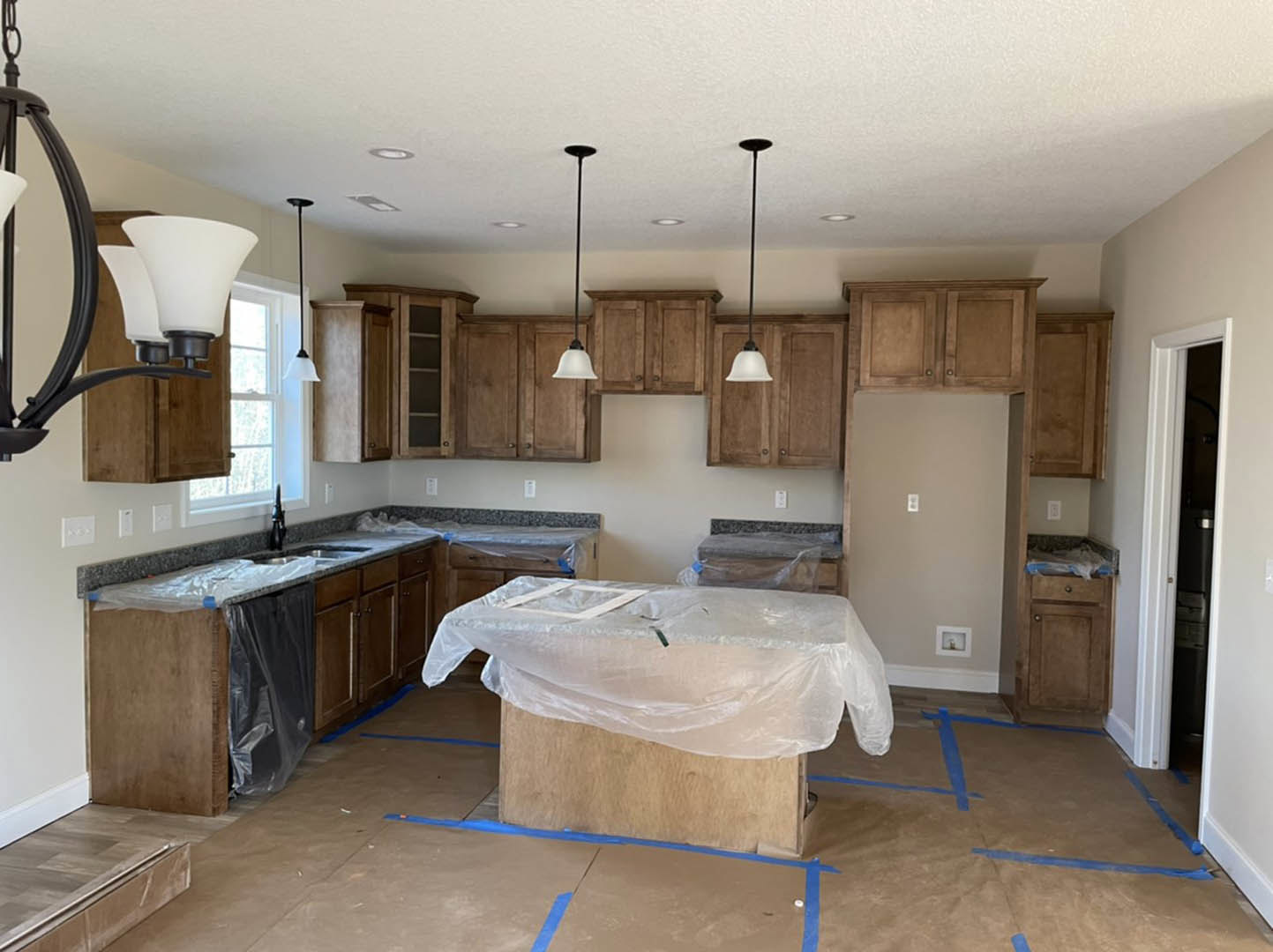 Kitchen with light wood cabinets, white countertops, stainless steel sink, and a dining table covered in clear plastic, set on light tile flooring
