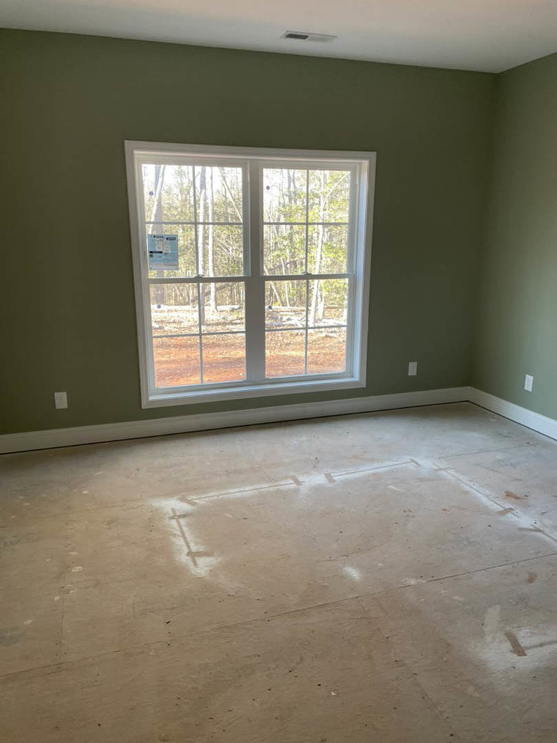 Concrete floor with visible water stains, green plastered wall, large window overlooking trees, natural daylight illuminating minimalist interior