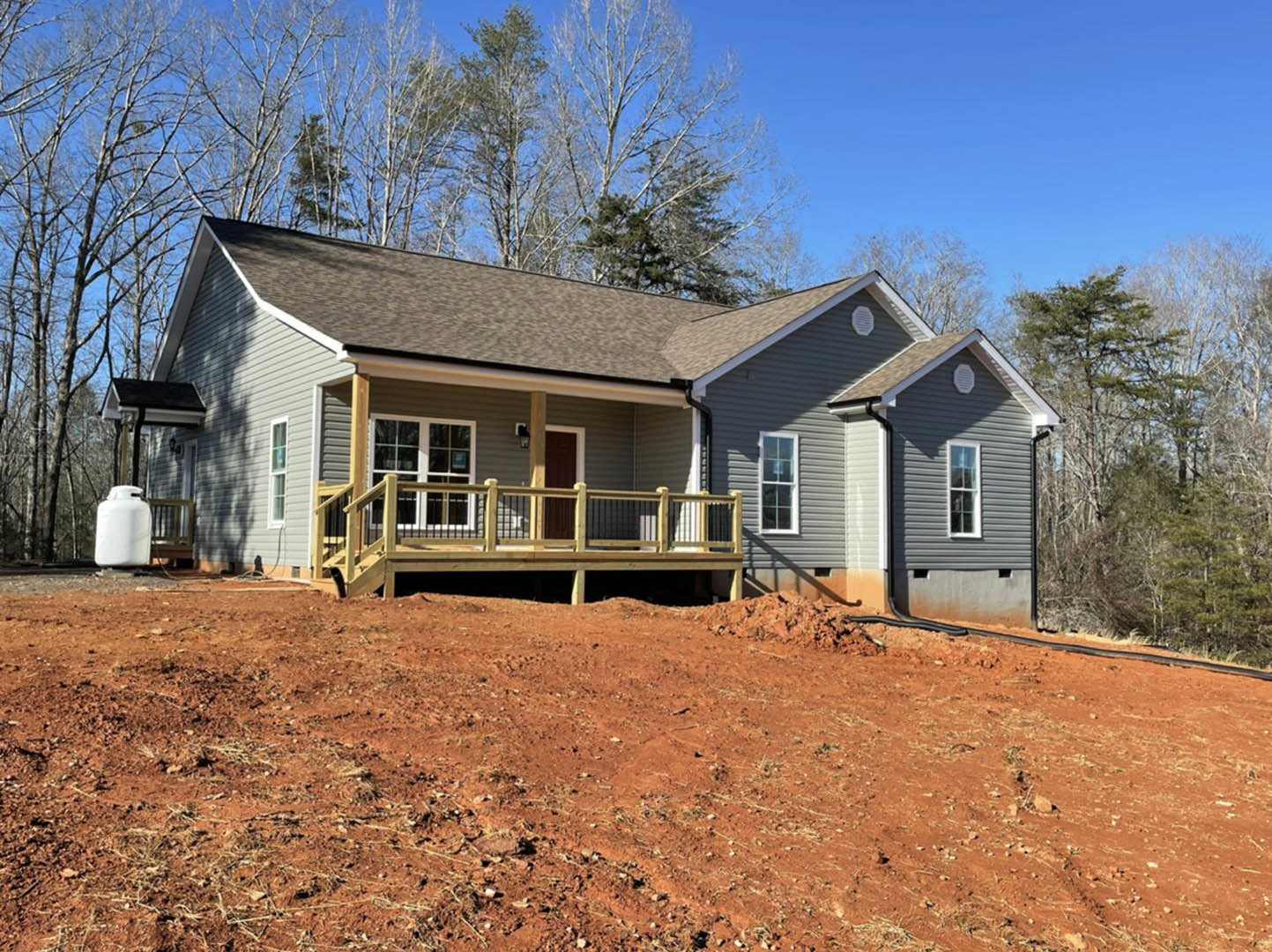 Two-story house with covered porch, wooden deck and railing, dirt area and pile in front, large windows, trees in background