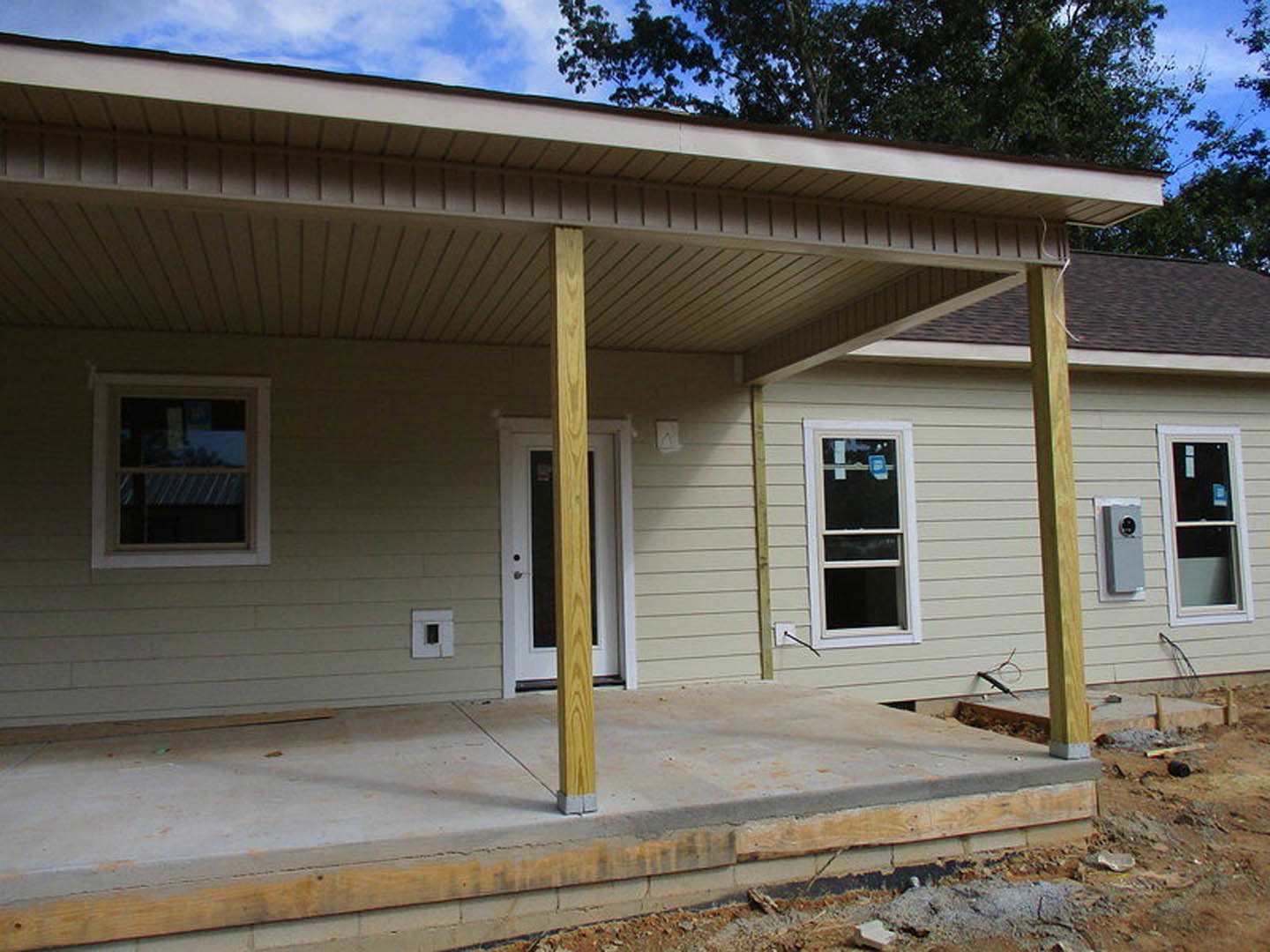 Partially built house with exposed wooden porch posts, concrete steps, multiple windows including one with a sign, light-colored siding, and tree reflections in glass.